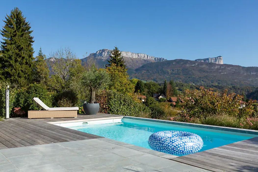 Outdoor pool with inflatable ring, sun lounger, mountain and trees in background.
